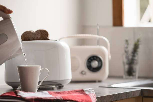 Hand pouring water for tea next to a white toaster with toast slices and a radio.