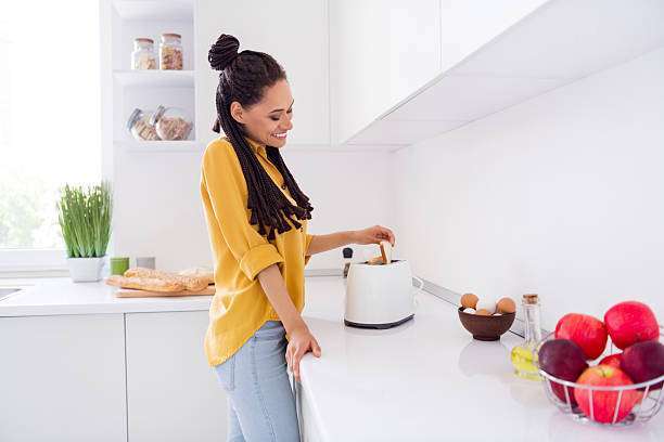 Smiling woman in yellow shirt making toast with a white toaster in a bright kitchen.